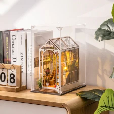 Miniature glass greenhouse inside a clear display case on a wooden shelf, warmly lit beside books and plants.