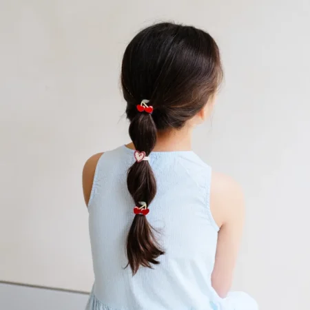 Back view of a young girl with a long braided ponytail secured by three red heart clips, wearing a light blue sleeveless dress.