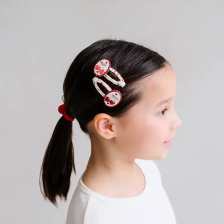 Side profile of a young girl with two white-and-red decorative hair clips in her dark hair, holding a ponytail with a red hair tie.