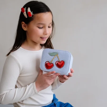 Young girl in a white sweater holds a small striped blue bag with red cherry embroidery.