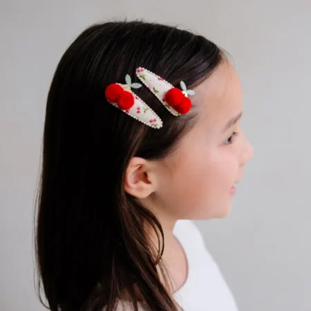 Side profile of a young girl wearing two white decorative hair clips with red pom-poms and small beads/flowers in her hair.