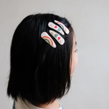 Back of a girl’s head with four decorative hair clips in rainbow and heart designs in dark hair, side view.
