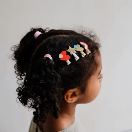 Side profile of a girl with curly hair and colorful heart and star hair clips on the side, including a rainbow clip.