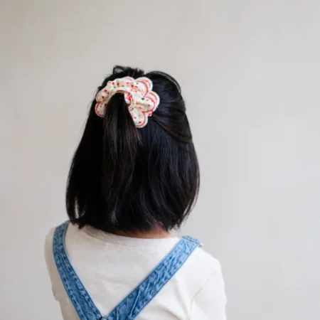 Back view of a person with short black hair wearing a white top and blue denim overalls, hair tied with a white polka‑dot scrunchie with red dots.