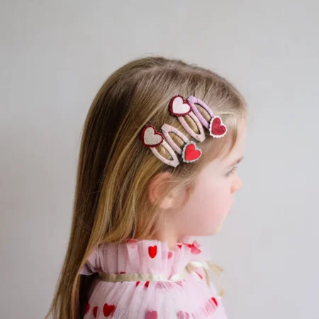 Side view of a young girl with pink and red heart hair clips arranged along her hairline, wearing a pink heart-patterned outfit.