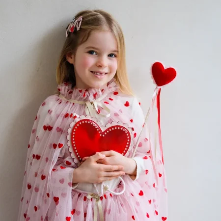 Young girl in a pink heart-pattern dress holding a red heart prop against a light wall.