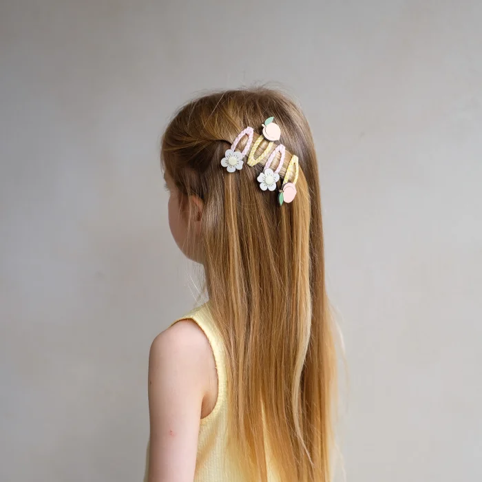 182098-23-peach-blossom-clic-clacs_69932c2a12b05_700x700r Side view of a girl with long light brown hair wearing pastel floral hair clips across the back, and a pale yellow top.