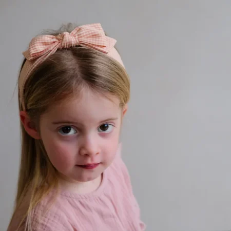 Young girl with light brown hair wearing a pink gingham bow headband, looking at the camera in a soft portrait.