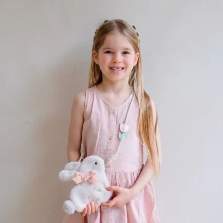 Smiling young girl in a pink dress holding a white plush bunny with a pink bow against a neutral wall.
