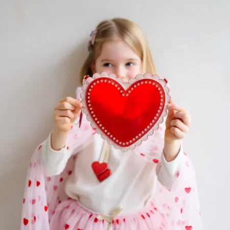 Young girl in a white sweater with pink hearts holding a red heart-shaped decoration toward the camera, for Valentine’s theme.