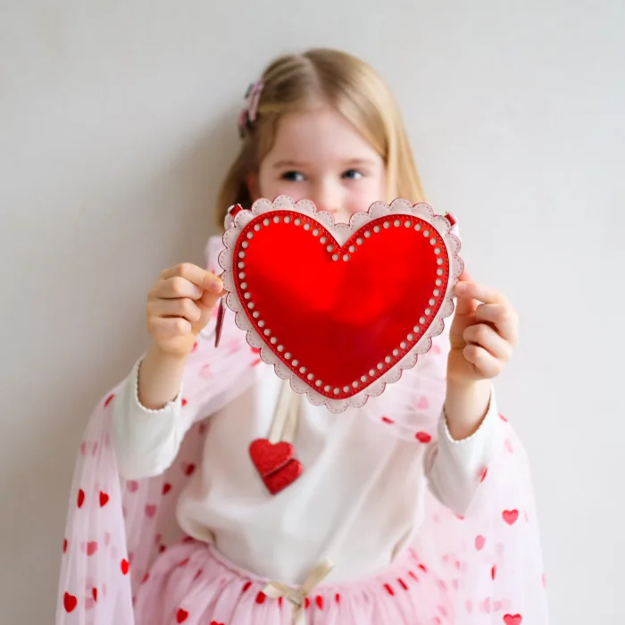 183003-14-scalloped-heart-bag-185002-27-heart-prin_69932a9ef2fed_700x700r Young girl in a white sweater with pink hearts holding a red heart-shaped decoration toward the camera, for Valentine’s theme.
