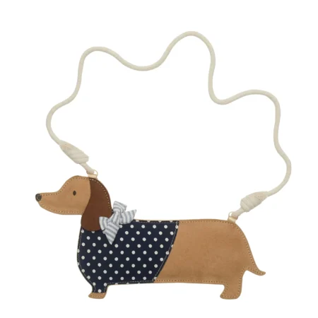 Dachshund-shaped crossbody bag with tan leather, navy polka-dot panel, bow, and a long cream strap.