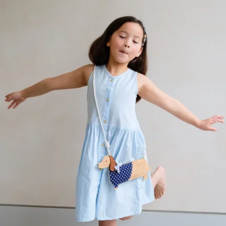Girl in a light blue sleeveless dress with arms outstretched, wearing a beige dog-shaped crossbody bag with blue polka-dot fabric.