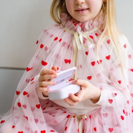 Young girl in a pink heart-pattern dress and sheer cape opening a small white jewelry box.