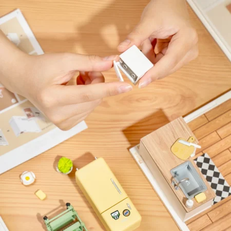 Hands assemble a small white mini cupboard part for a wooden toy kitchen on a wood floor.