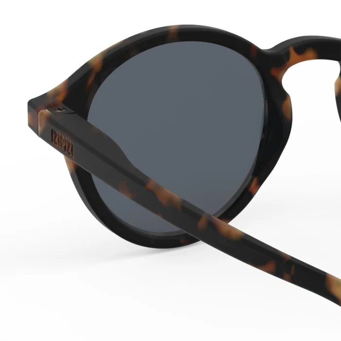 260_699daf5b558b8_700x700r Close-up of tortoiseshell sunglasses with dark gray lenses, showing the temple arm resting across the lens edge.
