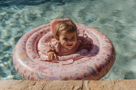 Baby girl in a pink floral pool floatation ring, smiling as she sits near the pool edge that sparkles in the water.