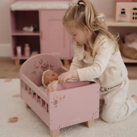 Little girl in a cream outfit kneels beside a pink wooden doll bed, tucking in a doll.