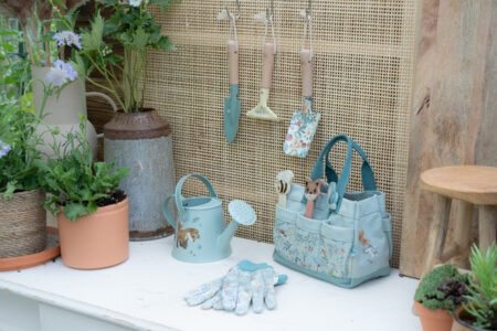 Pastel gardening setup on a white bench: hanging trowels, a watering can, gloves, and a blue tote bag nearby.