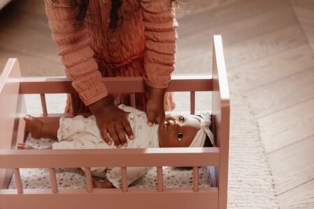 Young child in a pink dress leans over a pink crib, tending to a baby doll lying inside the crib.