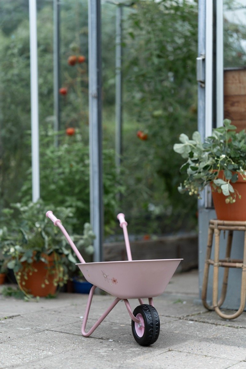 image_34552 Pink children's wheelbarrow with two pink handles and a single black wheel sits on a concrete path in a garden.