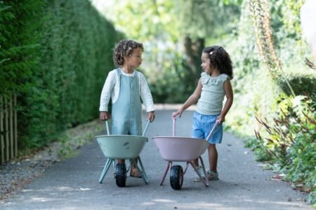 Two young children push pastel-colored toy wheelbarrows along a shaded garden path.