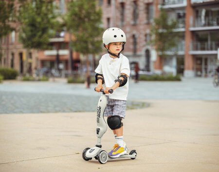 Child wearing a helmet and knee/elbow pads rides a white kick scooter in an outdoor plaza.