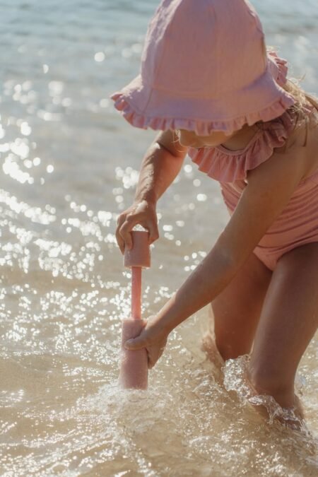 A young child in a pink swimsuit and sun hat kneels in shallow water, playing with a pink foam toy on the beach.
