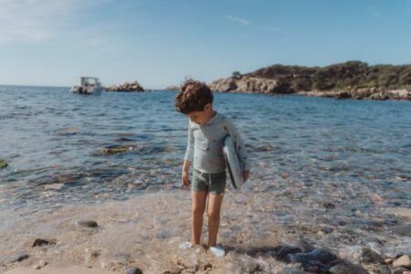 Boy standing in shallow water at a rocky beach, holding a blue surfboard under his arm as the sea and distant boat loom behind him.