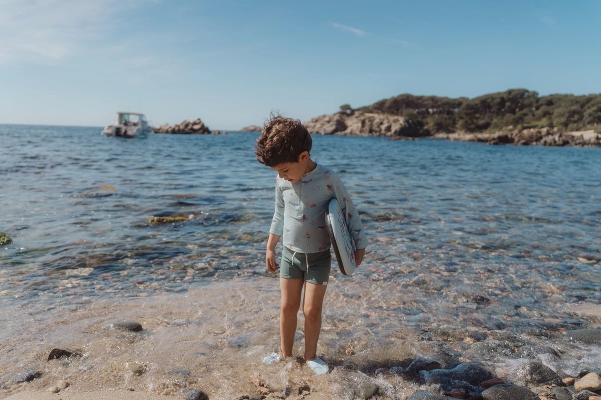 image_40604 Boy standing in shallow water at a rocky beach, holding a blue surfboard under his arm as the sea and distant boat loom behind him.