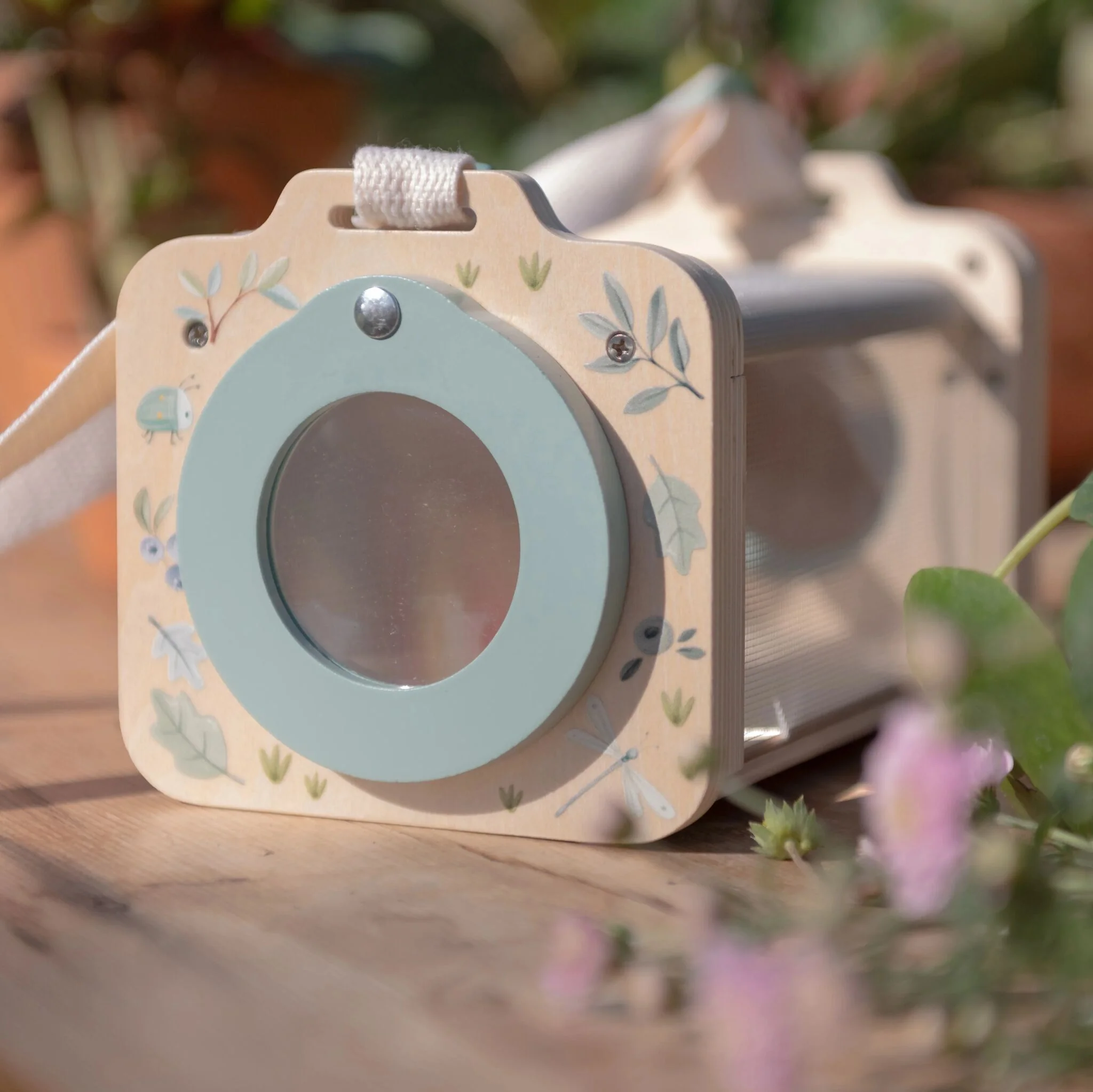 image_40802 Close-up of a wooden toy with a light blue circular inset and vine patterns, a white strap on top, with a blurred garden in the background.