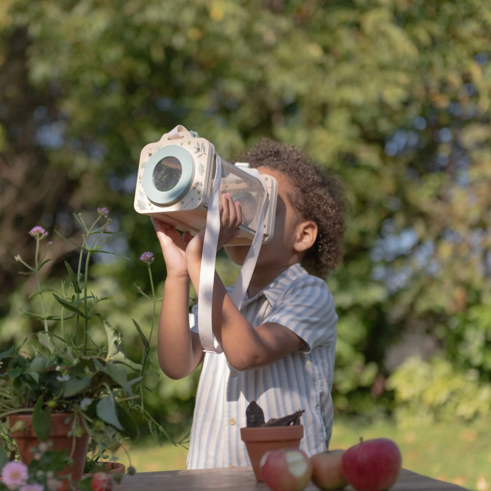 image_40803 Child outdoors wearing a vintage-style wooden camera on their head, aiming it toward the table with apples and plants around.