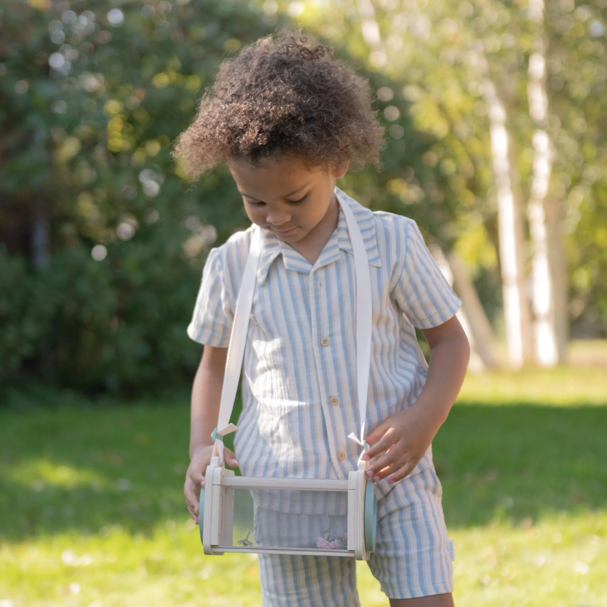 image_40807 Child in light blue and white striped outfit outdoors, holding a small wooden box with a mesh front.