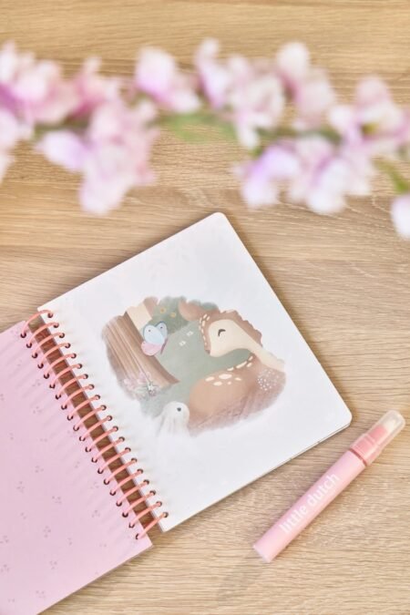 Pink spiral notebook with a dinosaur illustration on the page, placed on a wooden surface with a pink marker nearby and soft pink flowers blurred above.