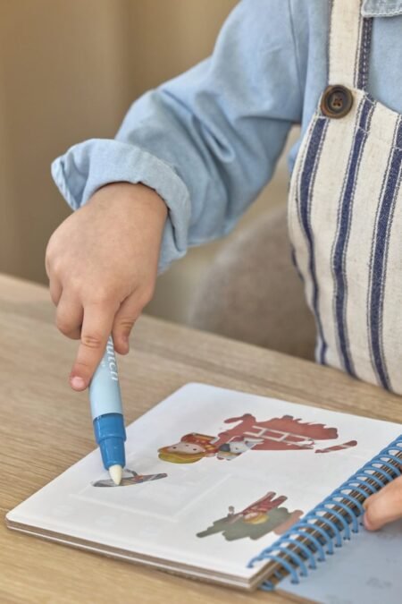 Child's hand with a blue marker coloring a cartoon page in a spiral notebook on a wooden table.