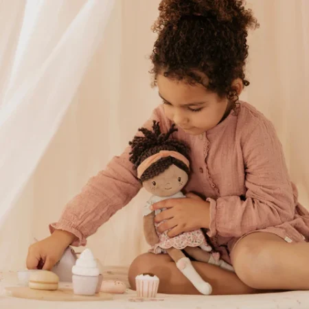 Young girl with curly hair in a pink dress sits cross‑legged, holding a doll while playing with toy pastries on a light surface.