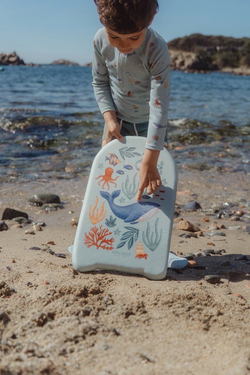 image_41428 Child on a sandy beach holding a light-blue sea-themed kickboard decorated with a whale, octopus, and coral near the water.