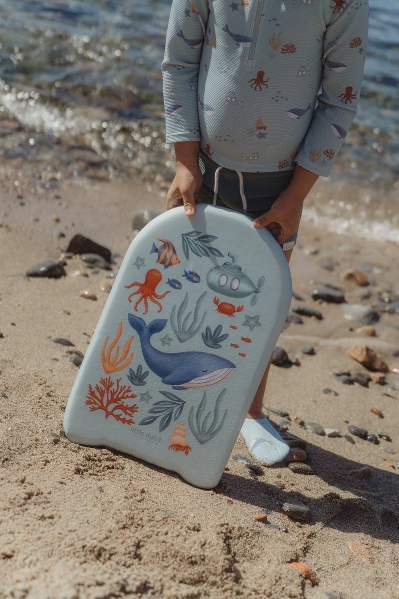 image_41429 Child on a sandy beach holding a light blue sea-themed kickboard with a whale, fish, octopus, and coral designs.