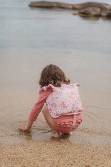 Young child crouching at the edge of the water on a sandy beach, touching wet sand with bare feet, wearing pink clothing and a patterned vest back view