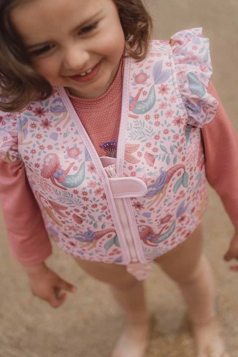 image_41437 Smiling young girl wearing a pink floral life vest standing on a sandy surface.