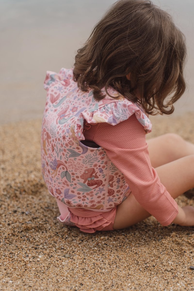 image_41438 Child sitting on a pebbled beach wearing a pink long-sleeve suit and a printed backpack with sea creatures, facing away from the camera