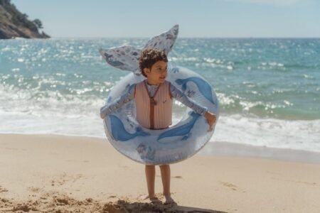 Young child in a striped life vest stands inside a blue whale-patterned inflatable ring on a sandy beach with the ocean in the background.