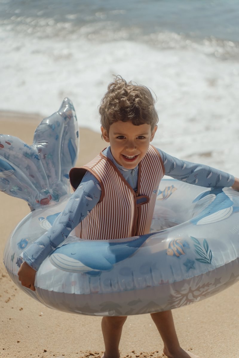 image_41442 Smiling boy in a striped life vest standing on the sandy beach with a blue inflatable swim ring around him near the water.