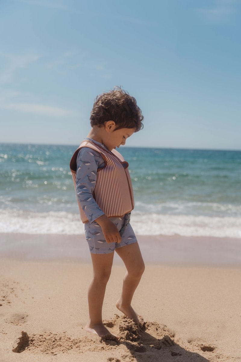 image_41443 Young child wearing a striped life jacket walks along the sandy beach toward the water with the ocean in the background.