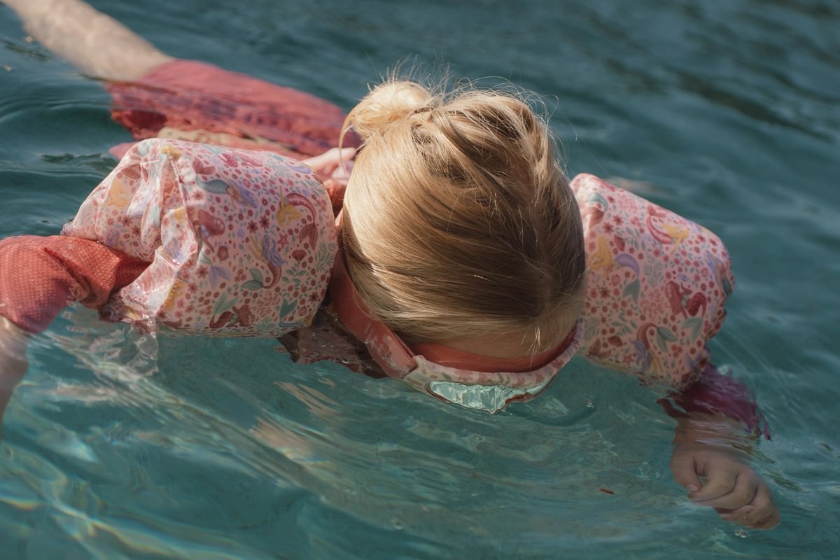image_41448 Child with blonde hair swimming on their stomach in clear blue water, wearing a pink floral swimsuit.
