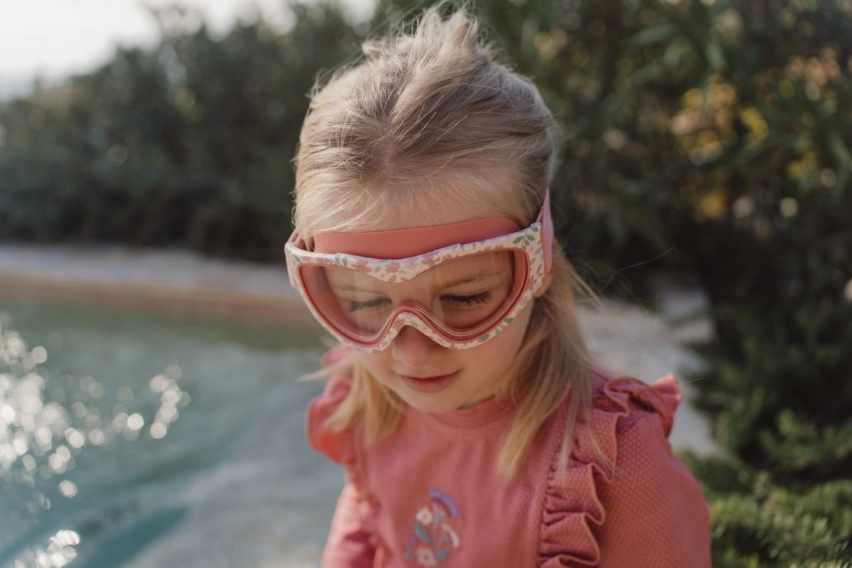 image_41449 Young girl wearing pink swimming goggles and a pink top by a pool, looking down thoughtfully.