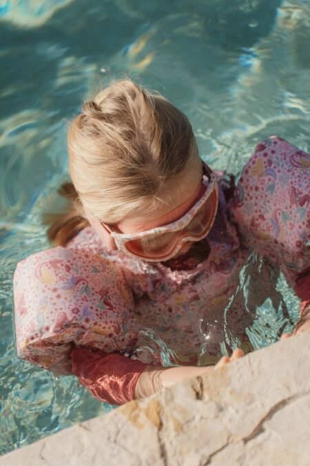 Girl in pink floral swimsuit and swim goggles sits at the edge of a pool, dipping into water as she climbs out.