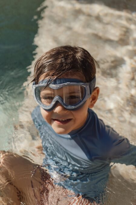Young boy wearing a snorkeling mask in clear water beside a rocky shore, smiling at the camera.