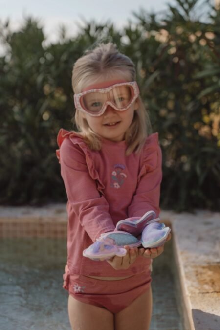 Young girl in a pink swimsuit and pink goggles stands by a pool, holding colorful water shoes.