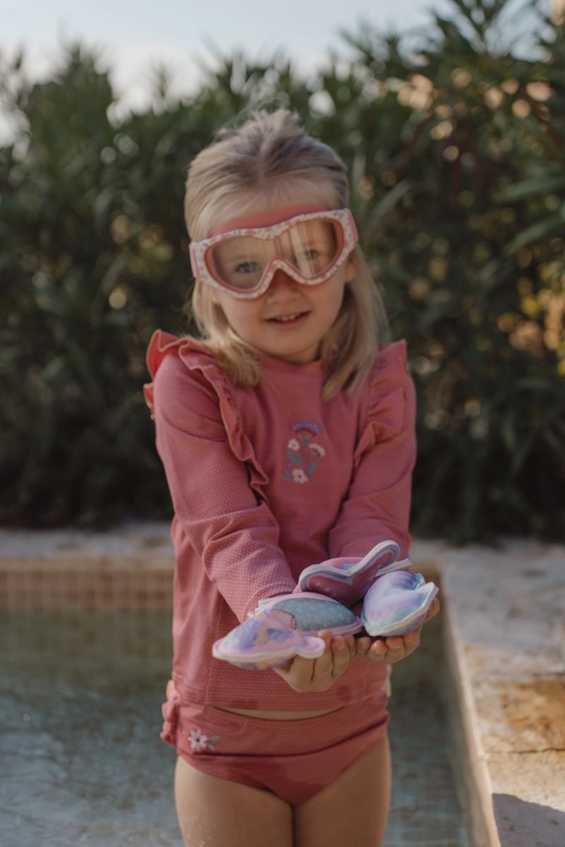 image_41631 Young girl in a pink swimsuit and pink goggles stands by a pool, holding colorful water shoes.